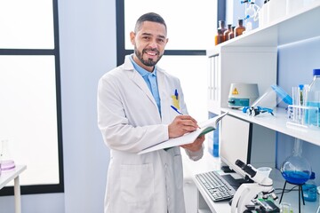 Young latin man scientist smiling confident writing on notebook at laboratory