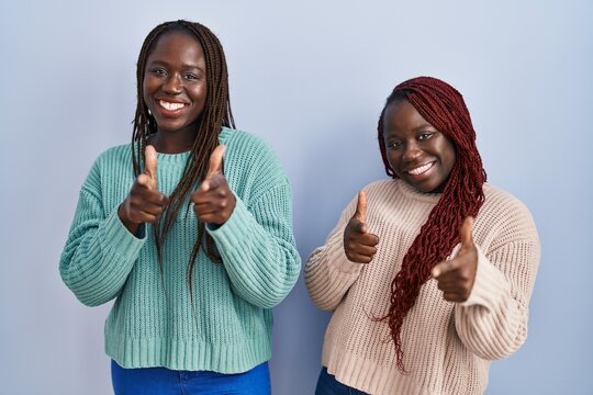 Two african woman standing over blue background pointing fingers to camera with happy and funny face. good energy and vibes.