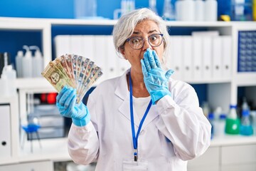 Middle age woman with grey hair working at scientist laboratory holding polish zloty banknotes covering mouth with hand, shocked and afraid for mistake. surprised expression