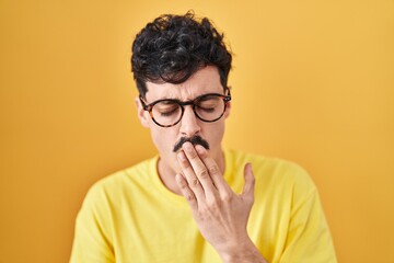 Hispanic man wearing glasses standing over yellow background bored yawning tired covering mouth with hand. restless and sleepiness.