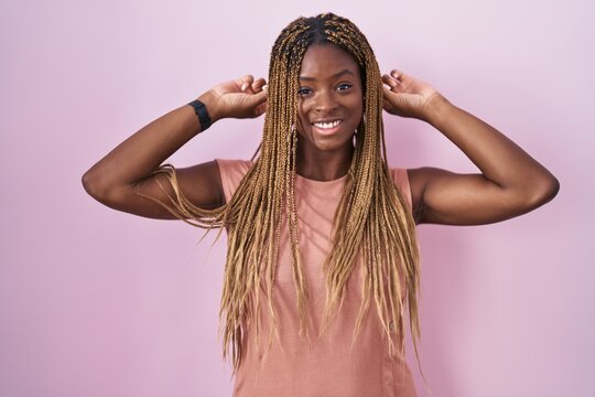 African american woman with braided hair standing over pink background smiling pulling ears with fingers, funny gesture. audition problem