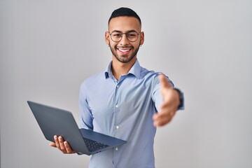 Young hispanic man working using computer laptop smiling friendly offering handshake as greeting and welcoming. successful business.