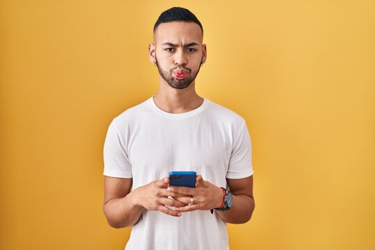 Young hispanic man using smartphone typing message puffing cheeks with funny face. mouth inflated with air, catching air.