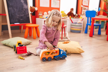 Adorable blonde girl playing with truck toy and dinosaur sitting on floor at kindergarten
