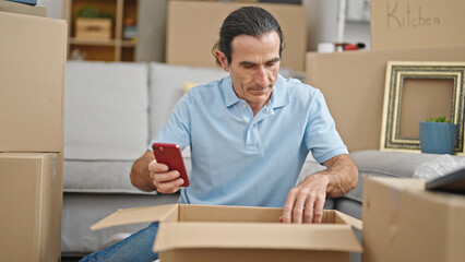 Middle age man unpacking cardboard box using smartphone at new home
