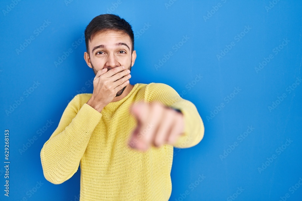 Poster Hispanic man standing over blue background laughing at you, pointing finger to the camera with hand over mouth, shame expression