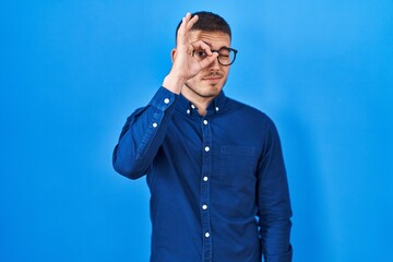 Young hispanic man wearing glasses over blue background doing ok gesture with hand smiling, eye looking through fingers with happy face.