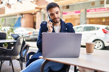 Young hispanic man talking on smartphone using laptop sitting on table at coffee shop terrace