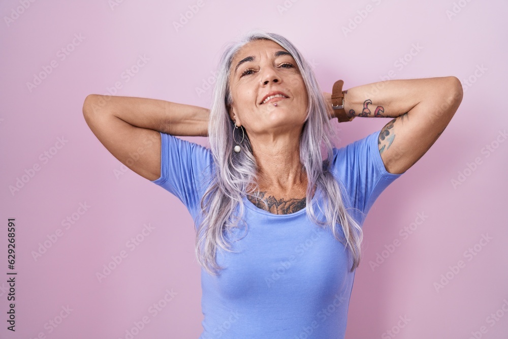 Wall mural Middle age woman with tattoos standing over pink background relaxing and stretching, arms and hands behind head and neck smiling happy