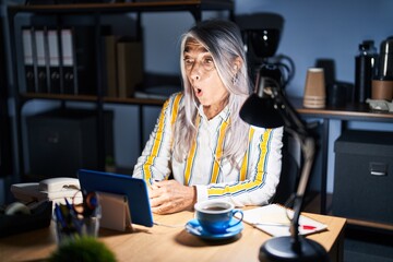 Middle age woman with grey hair working at the office at night afraid and shocked with surprise expression, fear and excited face.