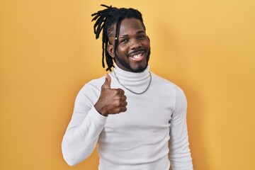 African man with dreadlocks wearing turtleneck sweater over yellow background doing happy thumbs up gesture with hand. approving expression looking at the camera showing success.