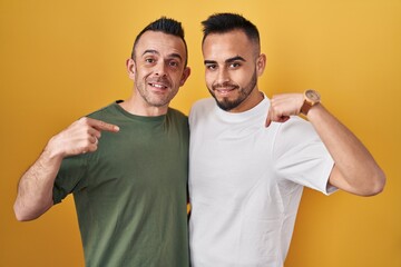 Homosexual couple standing over yellow background looking confident with smile on face, pointing oneself with fingers proud and happy.