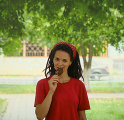 The concept of summer time joy. A young Caucasian woman is walking in the park and eating a chocolate popsicle on a stick. A girl with dreadlocks eats ice cream. Front view portrait.