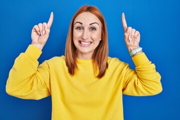 Young woman standing over blue background smiling amazed and surprised and pointing up with fingers and raised arms.