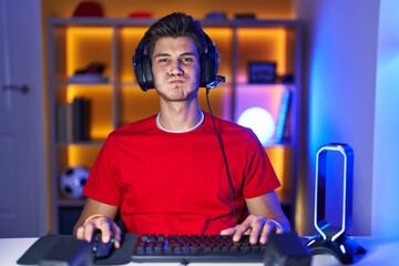 Young hispanic man playing video games puffing cheeks with funny face. mouth inflated with air,...