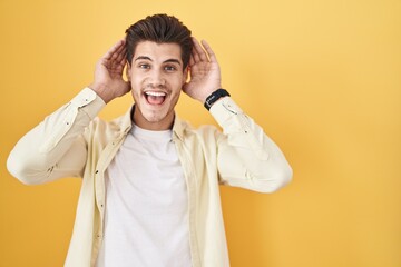 Young hispanic man standing over yellow background smiling cheerful playing peek a boo with hands showing face. surprised and exited