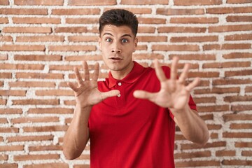 Young hispanic man standing over bricks wall afraid and terrified with fear expression stop gesture with hands, shouting in shock. panic concept.