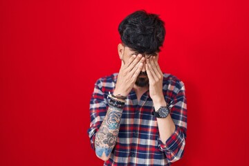 Young hispanic man with beard standing over red background rubbing eyes for fatigue and headache,...