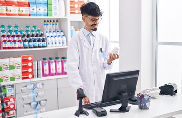 Young hispanic man pharmacist holding pills bottle using computer at pharmacy