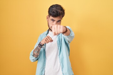 Young hispanic man with tattoos standing over yellow background punching fist to fight, aggressive and angry attack, threat and violence