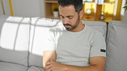 Young hispanic man sitting on sofa scratching arm at home
