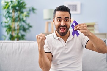 Young hispanic man with beard holding purple ribbon awareness screaming proud, celebrating victory and success very excited with raised arms