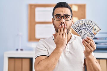 Young hispanic man with beard and glasses holding dollar banknotes covering mouth with hand,...