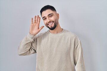 Young handsome man standing over isolated background waiving saying hello happy and smiling, friendly welcome gesture