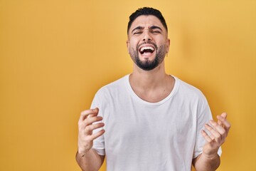 Young handsome man wearing casual t shirt over yellow background crazy and mad shouting and yelling with aggressive expression and arms raised. frustration concept.