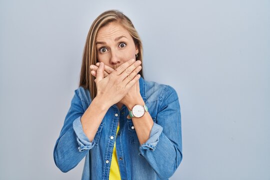 Young blonde woman standing over blue background shocked covering mouth with hands for mistake. secret concept.