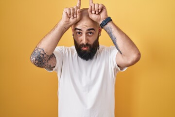 Young hispanic man with beard and tattoos standing over yellow background doing funny gesture with finger over head as bull horns