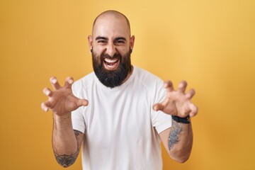 Young hispanic man with beard and tattoos standing over yellow background smiling funny doing claw...