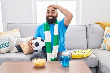 Young hispanic man with beard and tattoos football hooligan holding ball supporting team stressed...