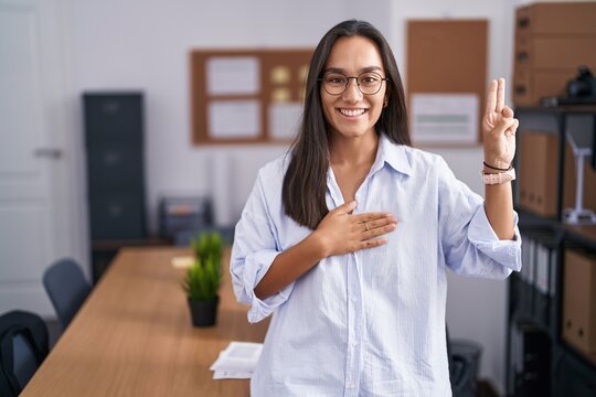 Young hispanic woman at the office smiling swearing with hand on chest and fingers up, making a loyalty promise oath