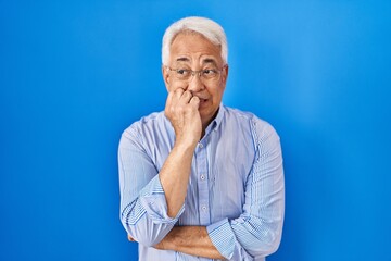 Hispanic senior man wearing glasses looking stressed and nervous with hands on mouth biting nails. anxiety problem.