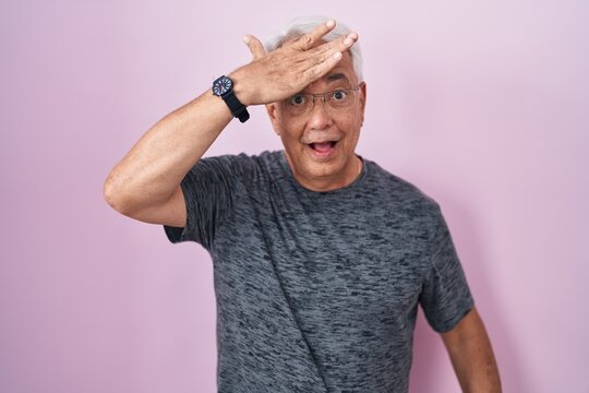 Middle age man with grey hair standing over pink background surprised with hand on head for mistake, remember error. forgot, bad memory concept.