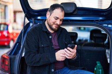 Young hispanic man using smartphone sitting on car trunk at street