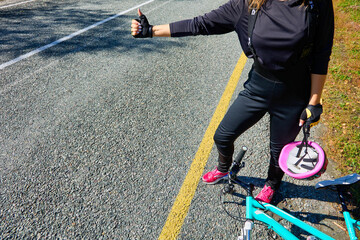 Woman in a tracksuit, gloves and helmet in hand votes on the road standing next to a broken bicycle lying down. Female cyclist votes with her hand to stop a car on an asphalt road