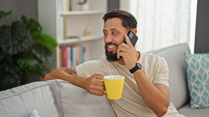 Young hispanic man talking on smartphone drinking coffee at home