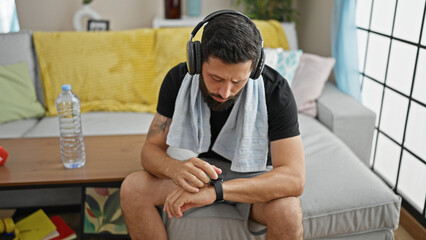 Young hispanic man sitting on sofa listening to music touching watch at home