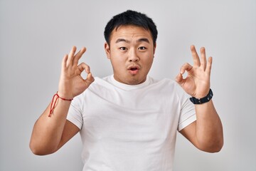 Fototapeta premium Young chinese man standing over white background looking surprised and shocked doing ok approval symbol with fingers. crazy expression