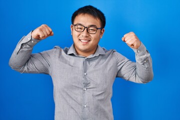 Young chinese man standing over blue background showing arms muscles smiling proud. fitness concept.