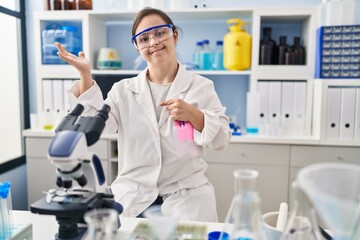 Hispanic girl with down syndrome working at scientist laboratory amazed and smiling to the camera while presenting with hand and pointing with finger.