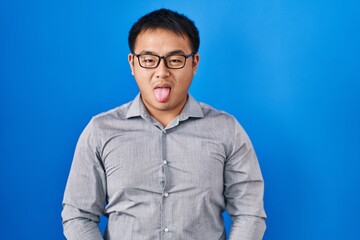 Young chinese man standing over blue background sticking tongue out happy with funny expression. emotion concept.