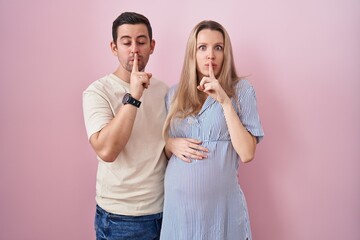 Young couple expecting a baby standing over pink background asking to be quiet with finger on lips....