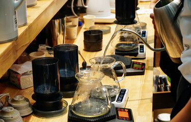 Hands of barista brewing a drip hot espresso by pouring hot water from stainless kettle into black cup with weighing machine and equipment on wooden counter.