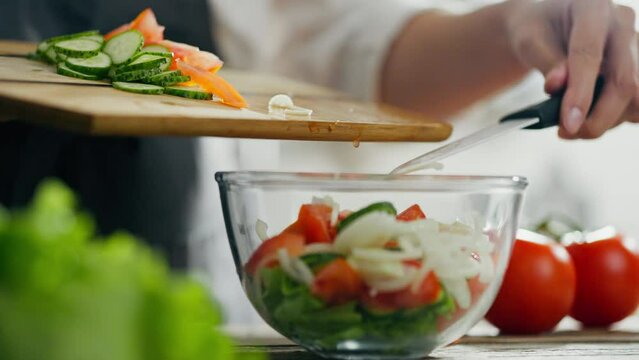 Female hands with knife cutting juicy vegetables on wooden cutting board to prepare a salad. Fresh vibrant summer Greek salad with tomats, onions, cucumbers in glass bowl. Concept of healthy lifestyle
