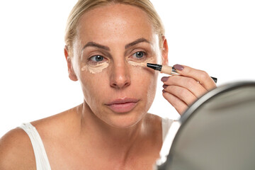 Middle aged senior woman applyes concealer under her eyes on white background