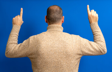 Portrait of a young man from behind, raising both hands and pointing the index finger up, wearing a beige pullover, isolated on a blue background