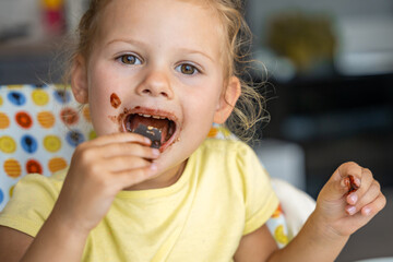 Little girl with blond hair eating homemade chocolate with dirty mouth and hands in home kitchen